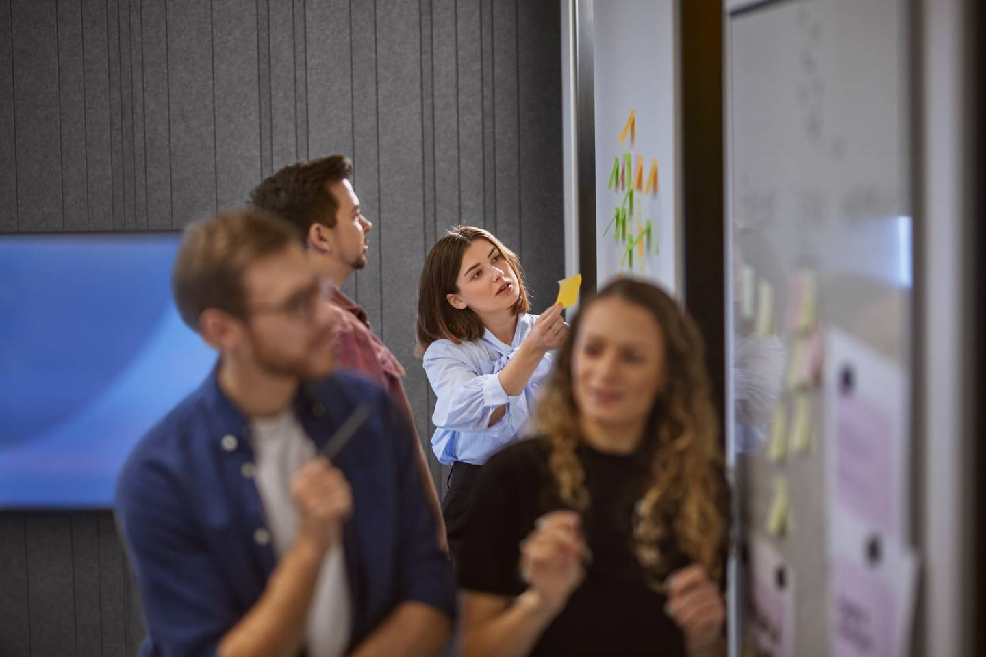 Kvinna sätter upp post-it på whiteboard under workshop med kollegor på kontor.