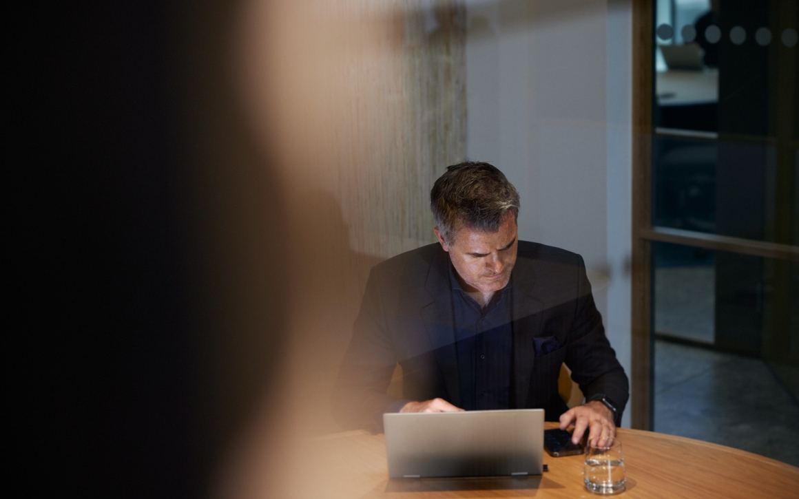 En man sitter vid ett runt bord med en laptop i kontorsmiljö.