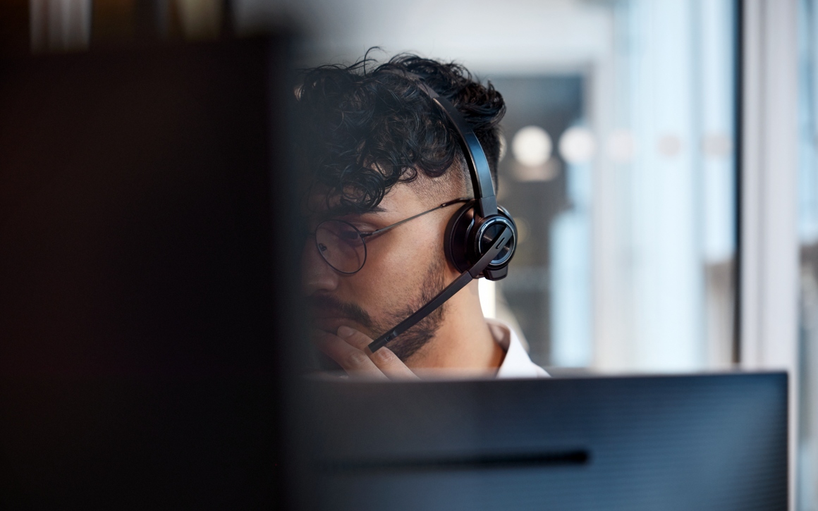 En man med ett headset på huvudet sitter framför datorskärmar i kontorsmiljö.